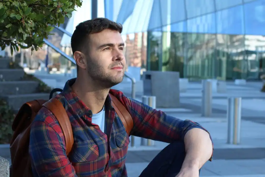 A man with short brown hair and a trimmed beard, wearing a plaid shirt and a brown backpack, sits outdoors near modern glass buildings, looking pensively into the distance.