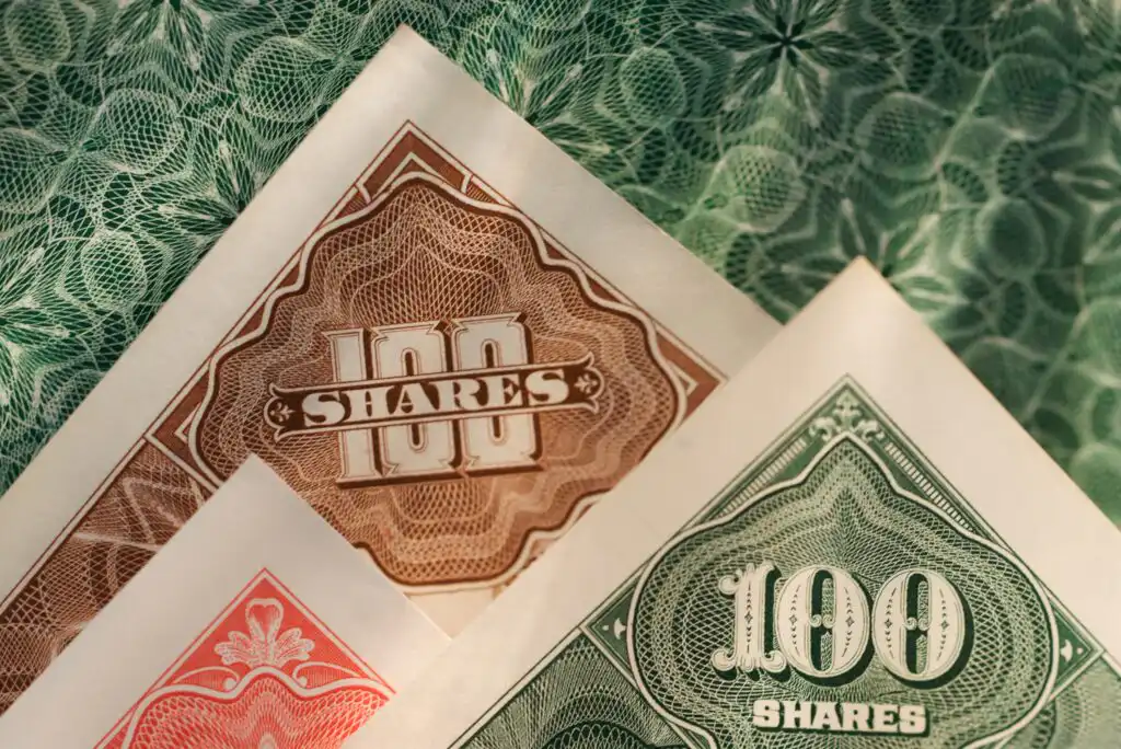 Close-up of three ornate stock certificates in brown, green, and red, each showing “100 SHARES” with intricate patterns and security designs in the background.