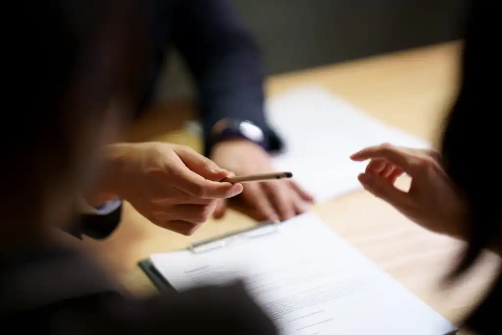 Two people sit at a table with documents; one hand is holding a pen and gesturing towards a clipboard with paperwork, while the other person reaches out, suggesting discussion or signing of documents.
