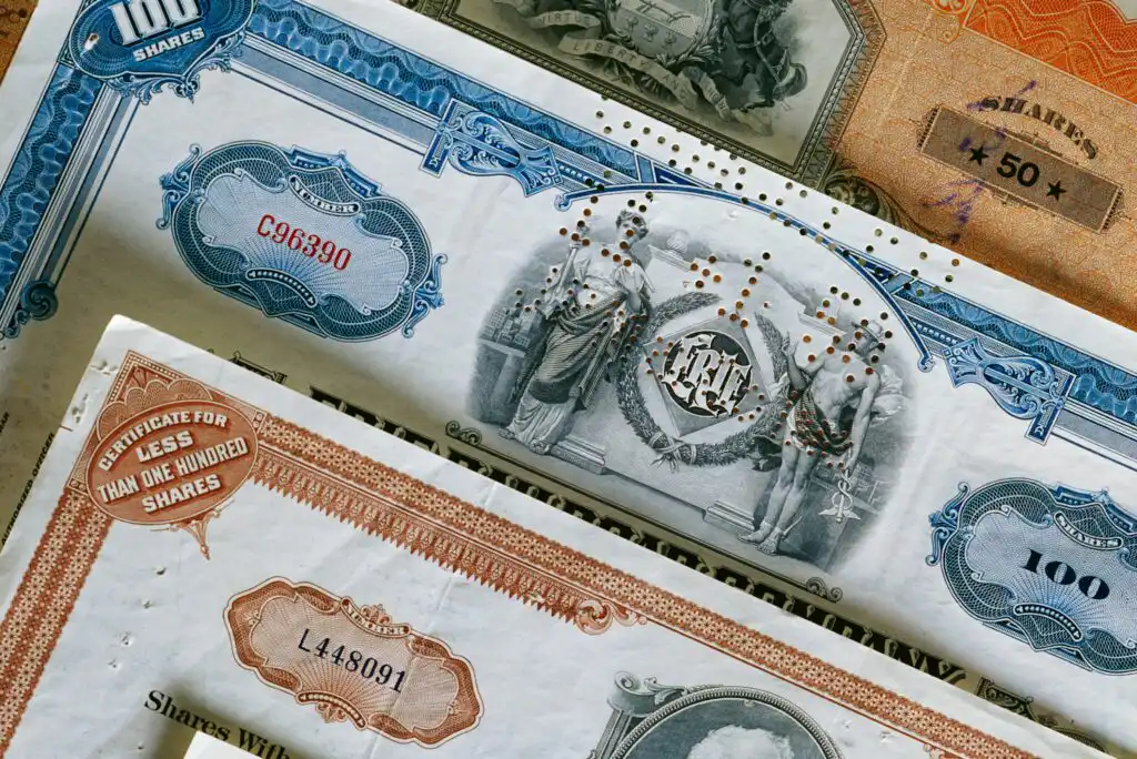 Close-up of vintage stock certificates in blue, orange, and white, featuring ornate borders, decorative designs, and official numbers, representing shares of different values and companies.