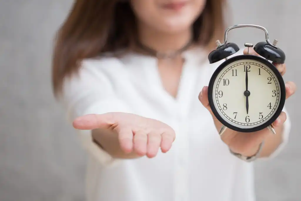 A woman in a white shirt holds a black analog alarm clock in one hand and extends her other open palm forward, with the clock showing the time as 8:00. Her face is partially out of focus.