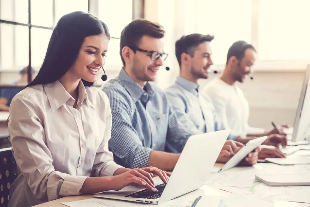 Four people wearing headsets sit in a row at a desk, working on laptops and tablets in a bright office, smiling and appearing engaged in customer support or a call center environment.