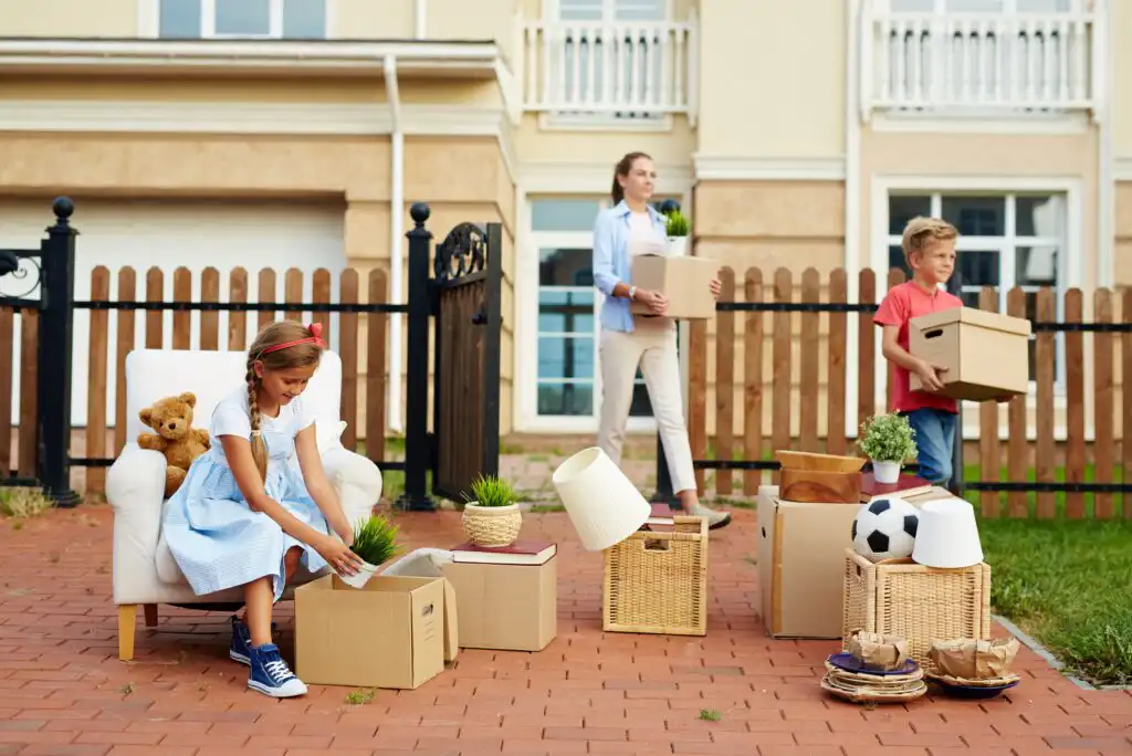 A young girl sits on a chair outside, unpacking a box, while a boy and a woman carry boxes in front of a house. Household items, plants, and a teddy bear are scattered around them on the patio.