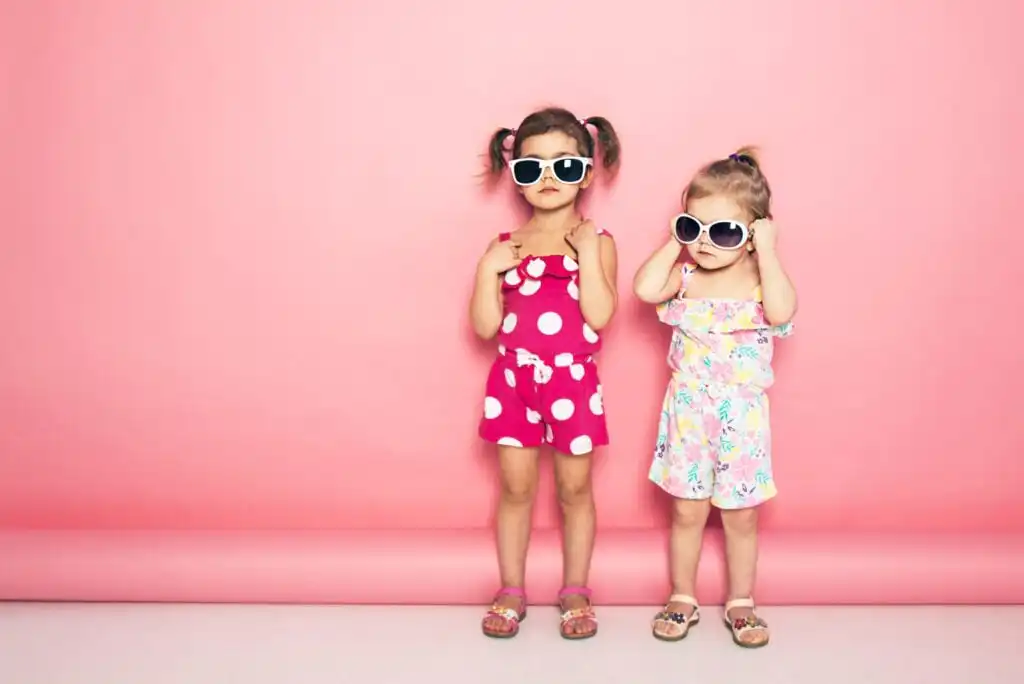 Two young girls wearing sunglasses stand against a pink background. One wears a pink polka dot romper, the other a colorful floral dress. Both have their hair in pigtails and are looking at the camera with serious expressions.