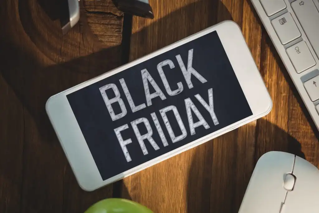 A smartphone on a wooden desk displays the words Black Friday in bold white letters. Nearby are a keyboard, a computer mouse, and part of an apple.