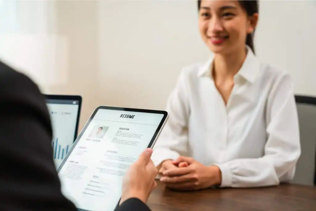 A person holds a tablet displaying a resume while interviewing a smiling woman in a white blouse, seated across a table in an office setting.