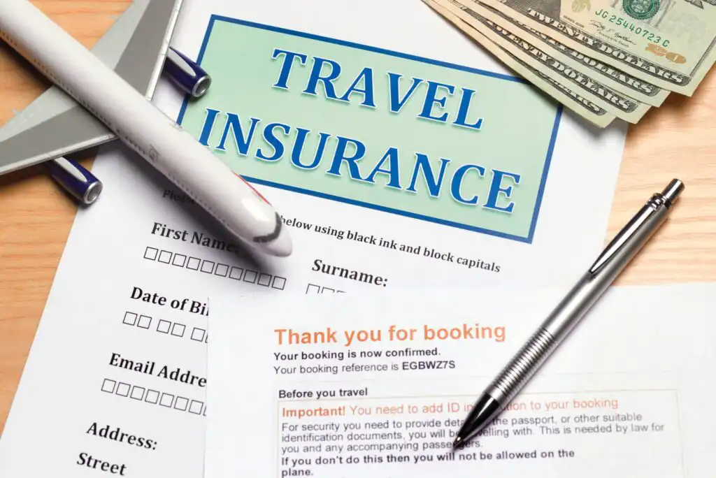 A travel insurance form sits on a desk with a toy airplane, pen, booking confirmation, and several U.S. dollar bills arranged around it.