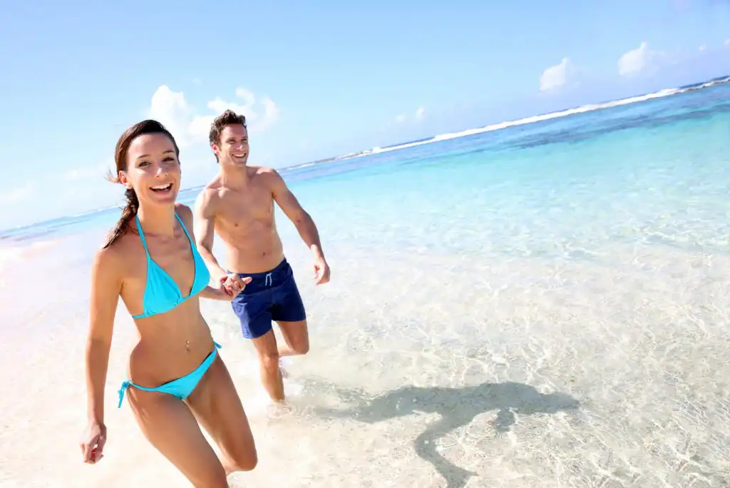 A smiling woman in a blue bikini and a man in blue swim trunks run through clear, shallow water at a sunny beach, with the ocean and blue sky in the background.