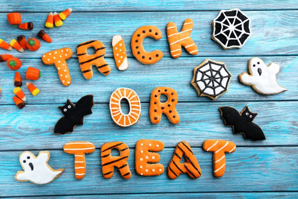Halloween cookies decorated as Trick or Treat, bats, ghosts, spider webs, and candy corn pieces arranged on a blue wooden background.