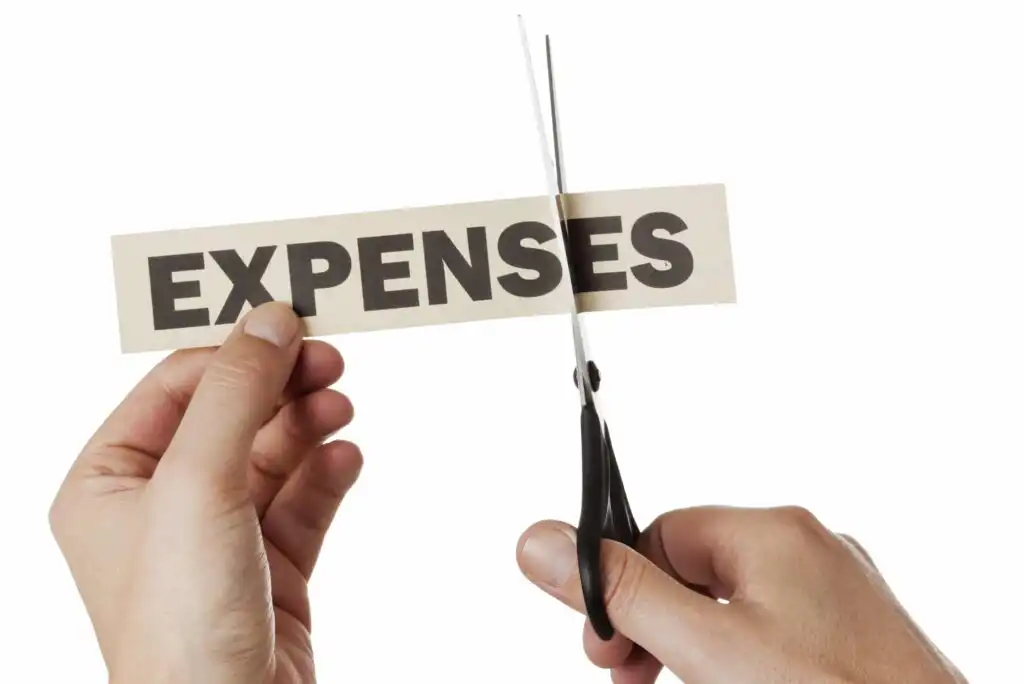 A person holds a paper with the word EXPENSES printed on it while another hand uses scissors to cut through the paper, symbolizing cutting expenses or reducing costs.