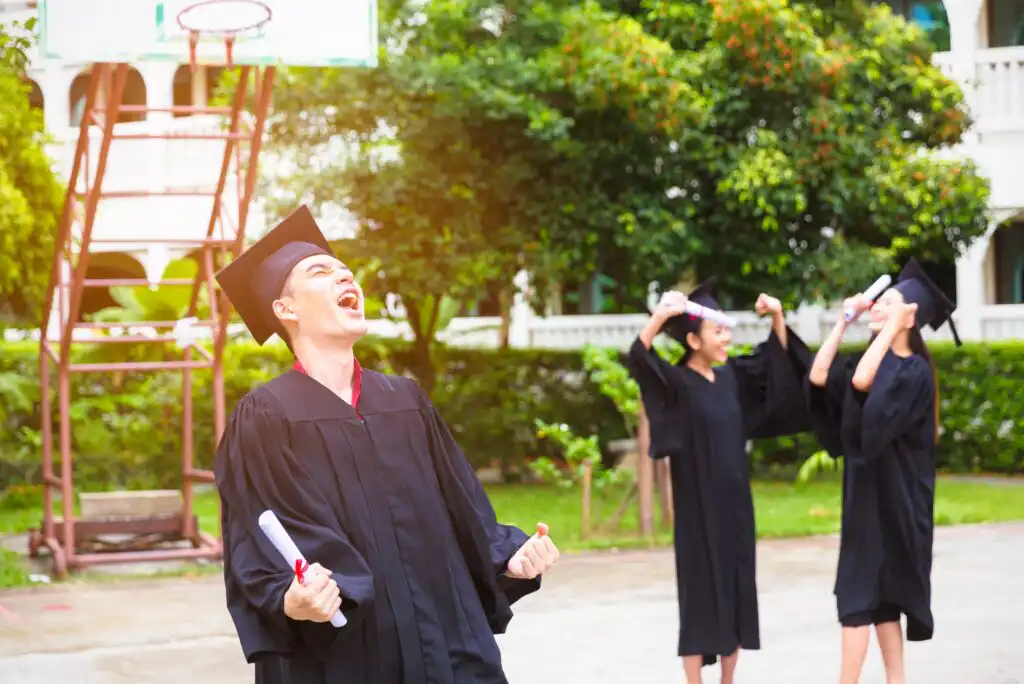 A group of graduates in black caps and gowns celebrate outdoors; one person in the foreground smiles and clenches their fists in excitement while two others rejoice in the background, holding diplomas.