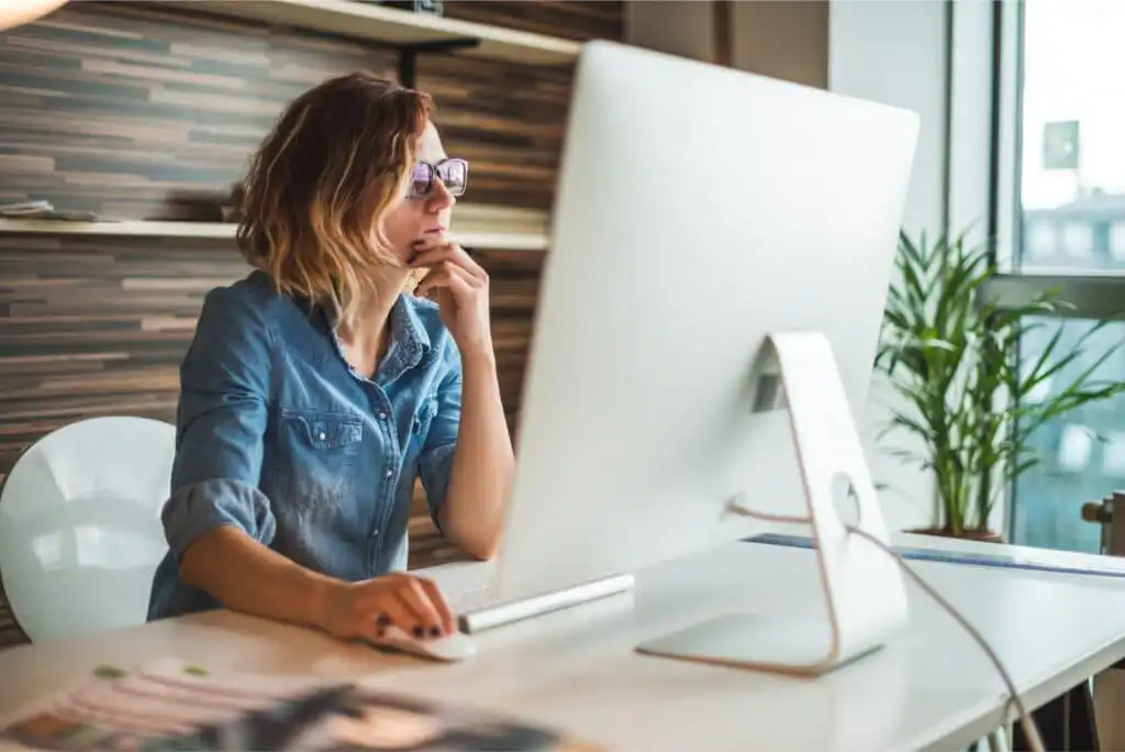 A woman wearing glasses and a denim shirt sits at a desk, looking thoughtfully at a large computer monitor in a modern office with wooden walls and a potted plant.