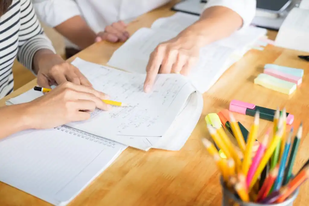 Two people studying together at a wooden desk with open notebooks, pens, highlighters, and pencils, as they write and point at notes and worksheets.