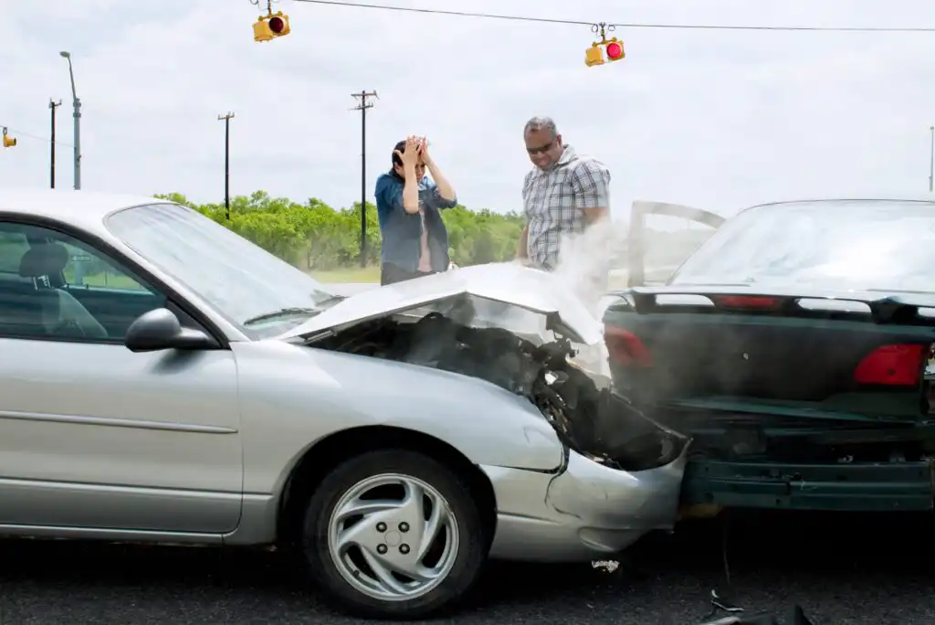 Two cars have collided at an intersection. A woman stands covering her face in distress, while a man inspects the damage. Smoke rises from the front of one car under cloudy skies and traffic lights.
