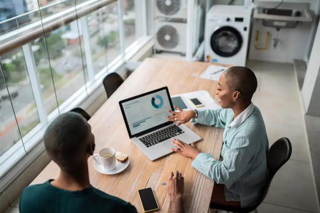 Two people sit at a wooden table in a bright room, looking at a laptop displaying graphs. One person gestures toward the screen while the other listens. A coffee cup, phone, and notebooks are on the table.
