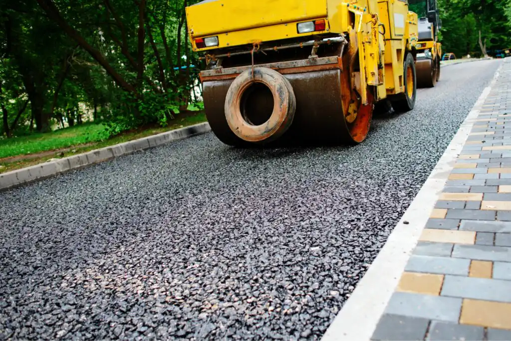 A yellow steamroller flattens fresh asphalt on a road, with a paved sidewalk and green trees visible alongside the road.