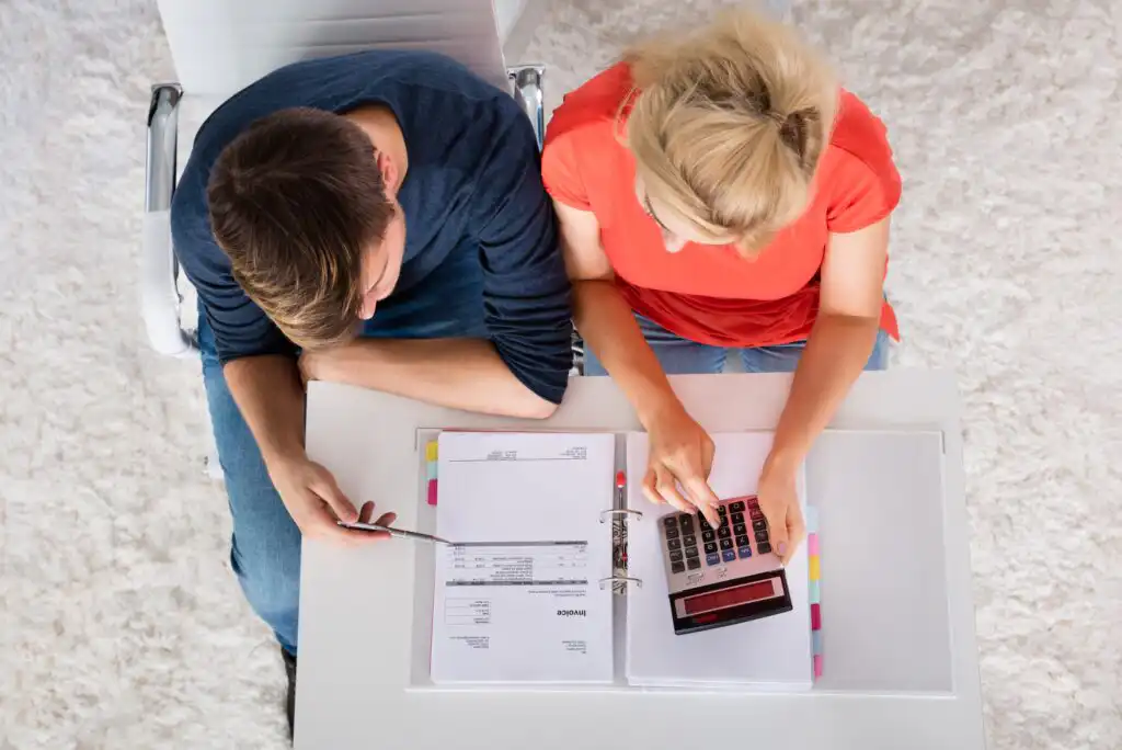 A man and woman sit at a white desk with financial documents, a calculator, and a binder, working together on budgeting or expenses. The photo is taken from above.