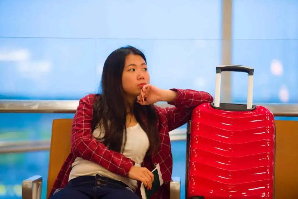 A woman sits in an airport waiting area next to a bright red suitcase, looking thoughtful and resting her chin on her hand. She is wearing a red plaid jacket and holding a phone and a passport.