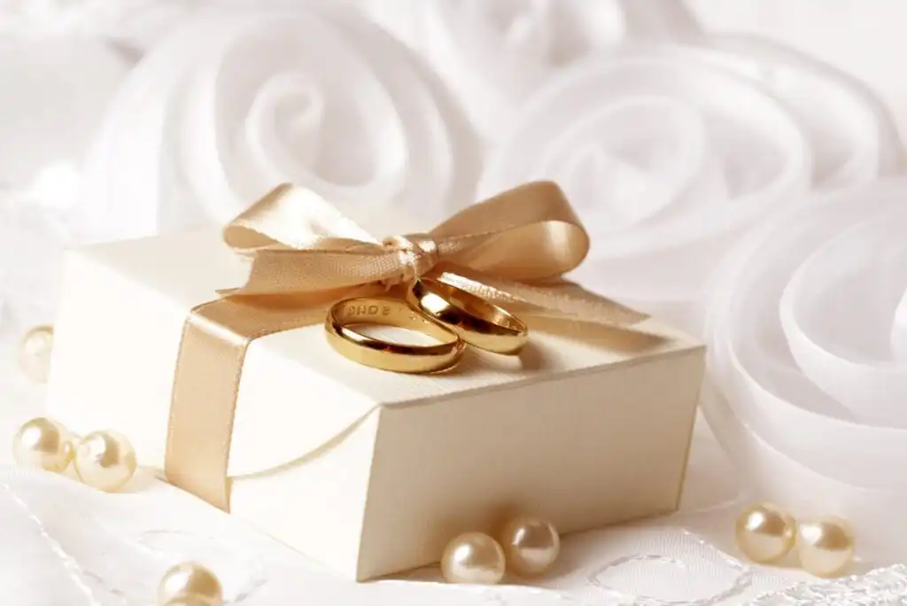 A cream-colored gift box with a gold satin ribbon bow, topped with two gold wedding bands, is surrounded by pearl beads and soft white fabric roses.