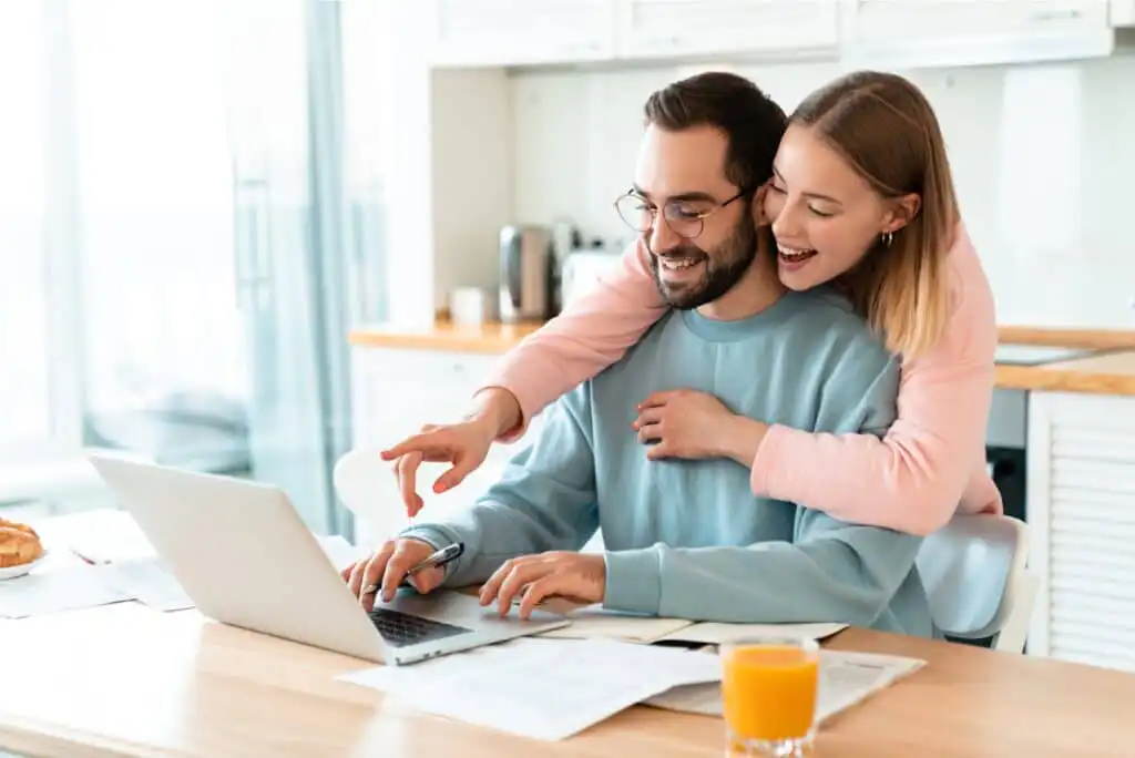 A smiling woman hugs a man from behind as he works on a laptop in a bright kitchen. Papers and a glass of orange juice are on the table, and the woman points at the screen, both appearing happy and engaged.