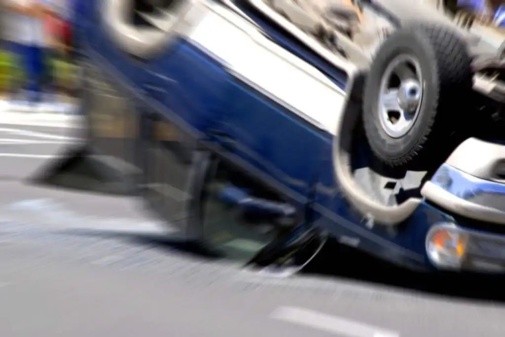 A blue car flipped upside down on a city street, with motion blur suggesting a recent crash. The vehicles undercarriage and wheels are visible, and debris is scattered on the road.