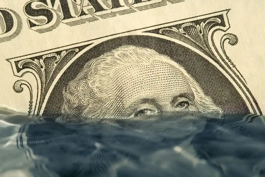 A close-up of a U.S. dollar bill with George Washingtons face partially submerged in water, symbolizing financial crisis or economic instability.