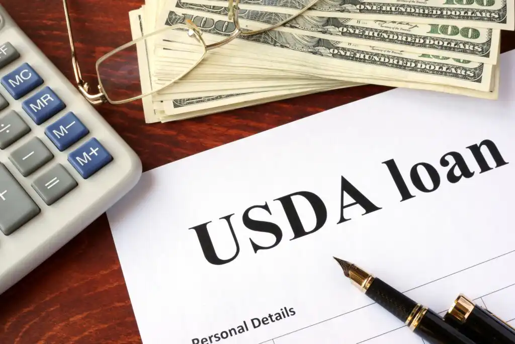 A document titled USDA loan sits on a desk next to a pen, a calculator, eyeglasses, and a stack of hundred-dollar bills.
