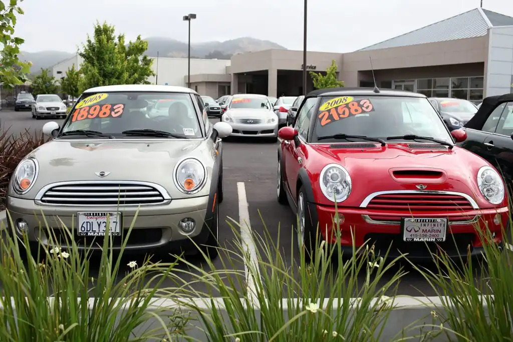 Two Mini Cooper cars, one silver and one red, are parked side by side at a car dealership. Price and year signs are displayed on the windshields, with other cars and a building in the background. Green plants are in the foreground.