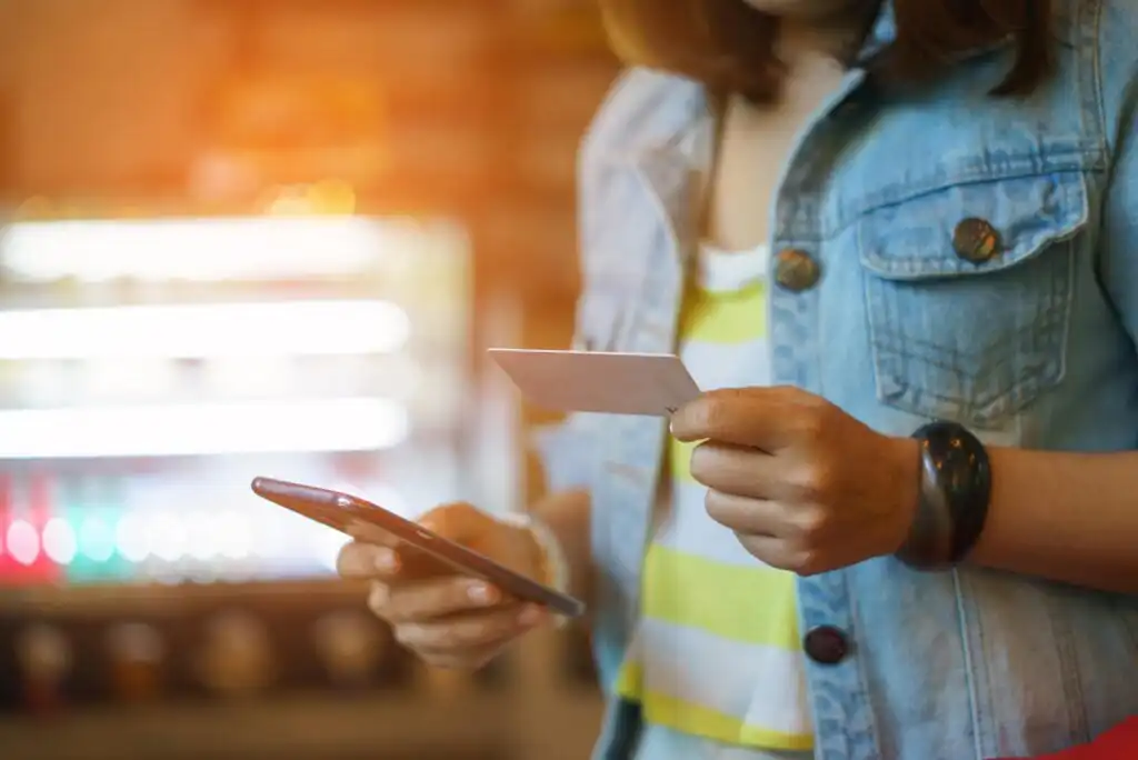 A person wearing a denim vest holds a credit card in one hand and uses a smartphone with the other, possibly making an online purchase or banking transaction, with blurred lights in the background.
