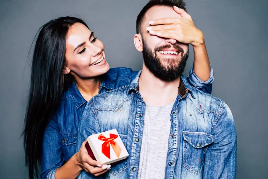 A woman smiles while covering a mans eyes with one hand and holding a small gift box with a red ribbon in the other. Both are wearing denim shirts and standing against a plain gray background.