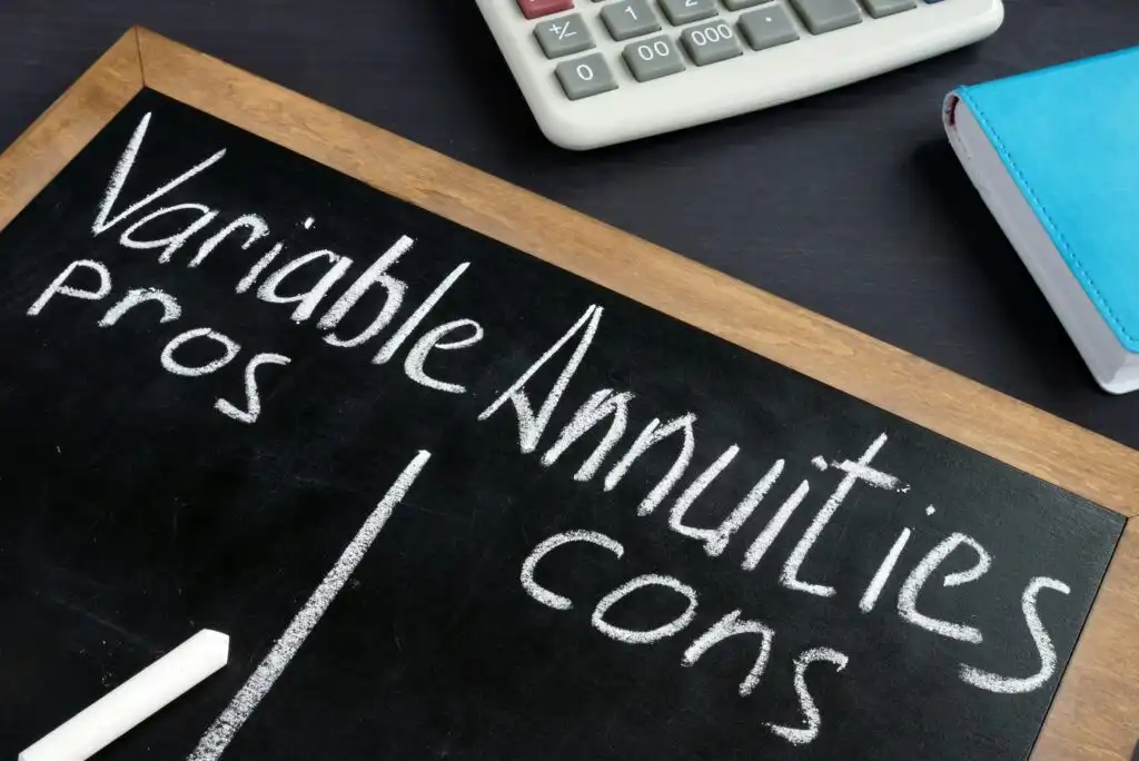 A chalkboard divided in half with “Variable Annuities” written at the top and “pros” on the left, “cons” on the right. A chalk piece, a calculator, and a blue notebook are nearby.