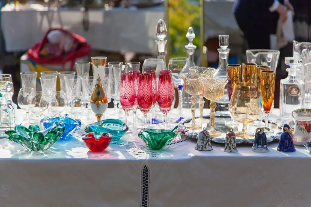 A table covered with a white cloth displays assorted colorful glassware, including goblets, bowls, vases, and figurines, at an outdoor market or fair in bright sunlight.