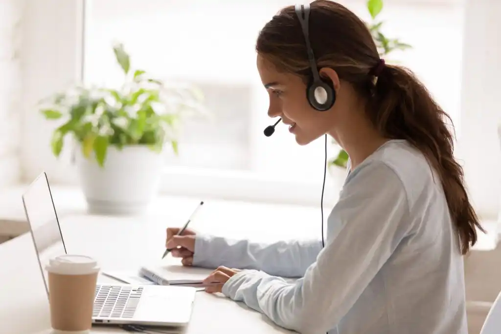 A young woman wearing a headset sits at a desk with a laptop, writing in a notebook. A coffee cup and potted plants are on the desk. She appears to be engaged in a virtual meeting or online class.