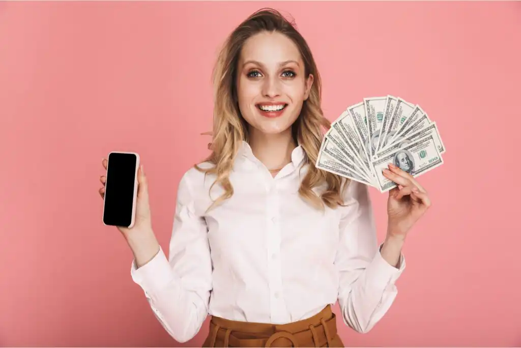 Smiling woman in a white shirt holding a smartphone in one hand and fanned out U.S. dollar bills in the other, standing in front of a pink background.