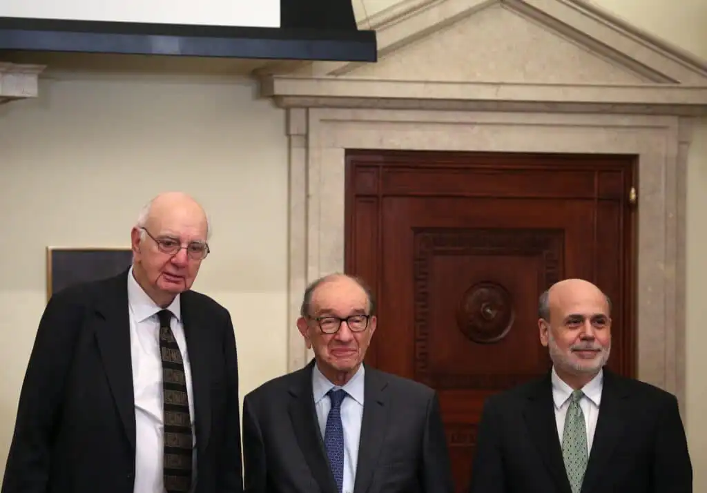 Three older men in suits stand side by side indoors in front of a wooden door with decorative trim and a stone arch above it. All three are looking at the camera and smiling slightly.