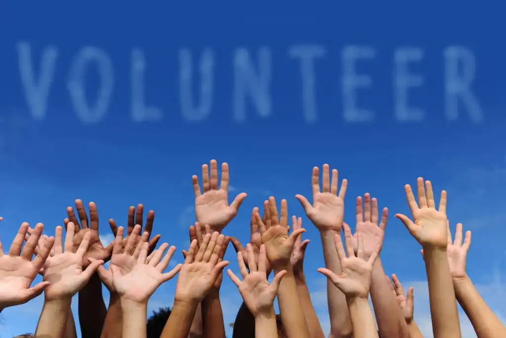 A group of raised hands of diverse people reaching up under a clear blue sky, with the word VOLUNTEER spelled out in white, cloud-like letters above them.