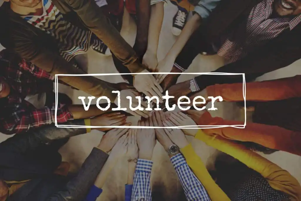 A group of diverse people standing in a circle place their hands together in the center. The word volunteer is written across the image in bold white letters.