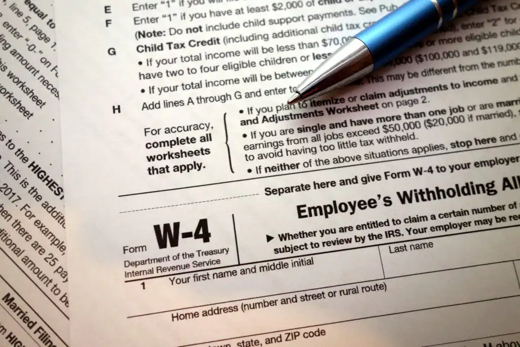 A blue pen rests on a U.S. IRS Form W-4 Employee’s Withholding Allowance Certificate, with sections of the form and instructions visible in the background.