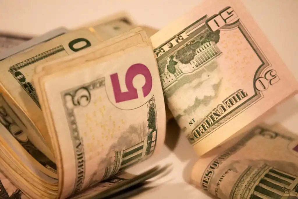A close-up view of rolled and stacked US dollar bills, including denominations of $5, $10, $20, and $50, arranged on a light surface.