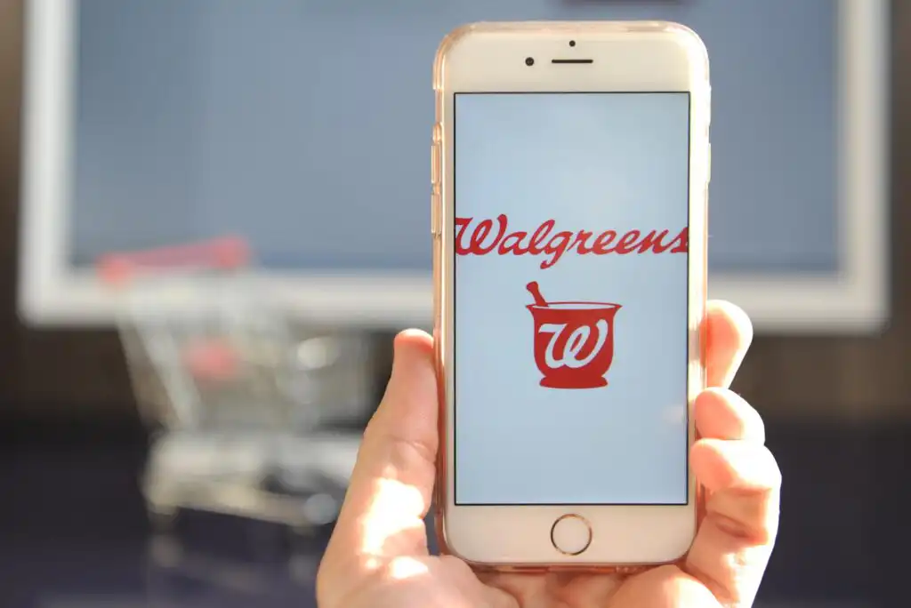 A hand holds a smartphone displaying the Walgreens logo and name in red on the screen. In the blurred background, a shopping cart is visible indoors.