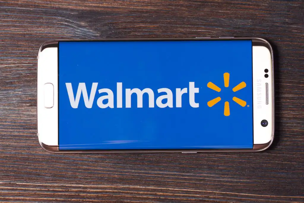 A smartphone lying on a wooden surface displays the blue Walmart logo and bright yellow spark symbol on its screen.