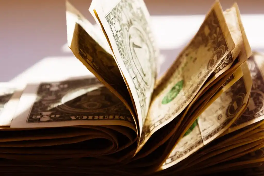 Several folded U.S. one-dollar bills are stacked and fanned out, with sunlight casting shadows and highlighting the texture and details of the paper currency.