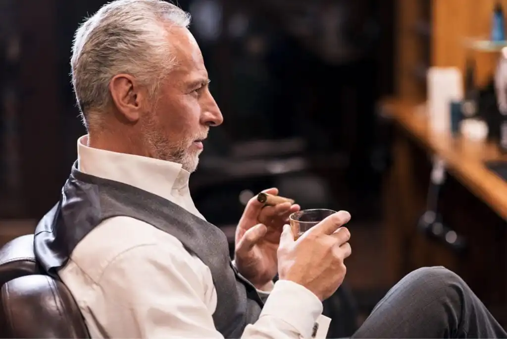 An older man with gray hair and a beard sits in a chair, wearing a white dress shirt and gray vest. He holds a cigar in one hand and a glass of whiskey in the other, looking contemplative.