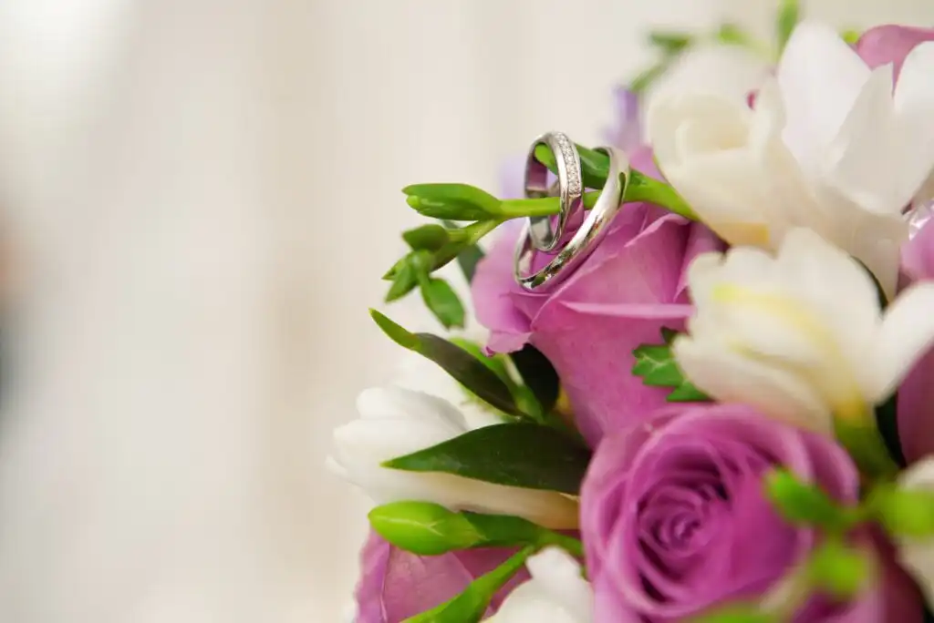 Two silver wedding rings rest on a bouquet of purple and white flowers, with green leaves and soft, blurred background.