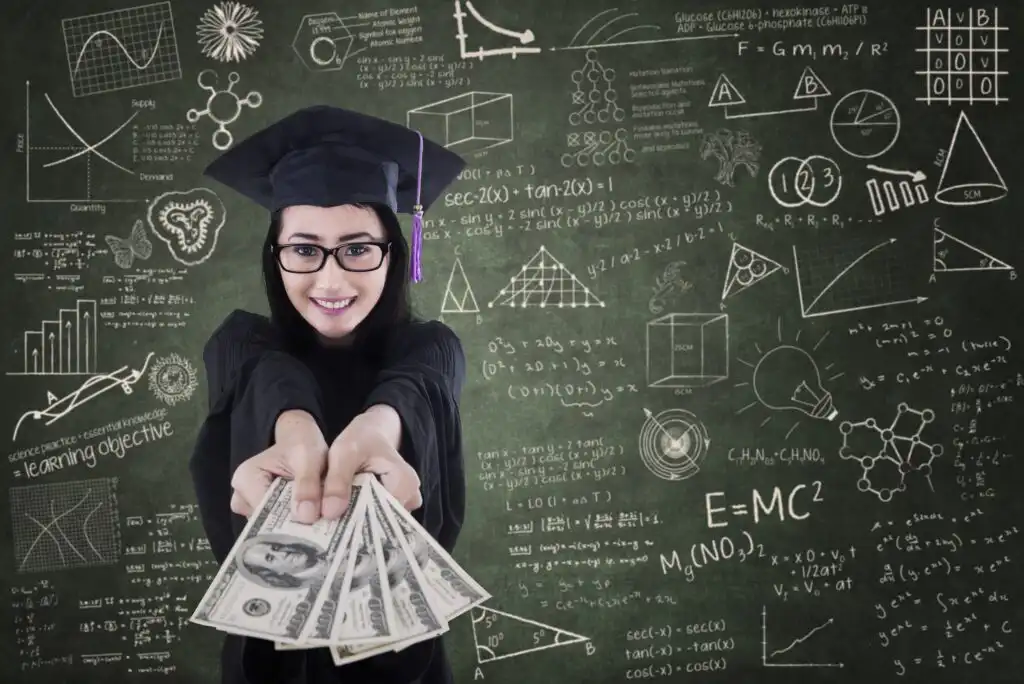 A smiling graduate in cap and gown holds out several $100 bills, standing in front of a chalkboard covered with scientific formulas, diagrams, and math equations.