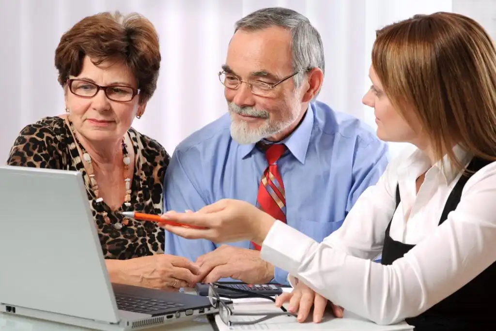 Three adults sit at a desk looking at a laptop. A younger woman in business attire gestures toward the screen while an older man and woman listen attentively, suggesting a discussion or consultation.