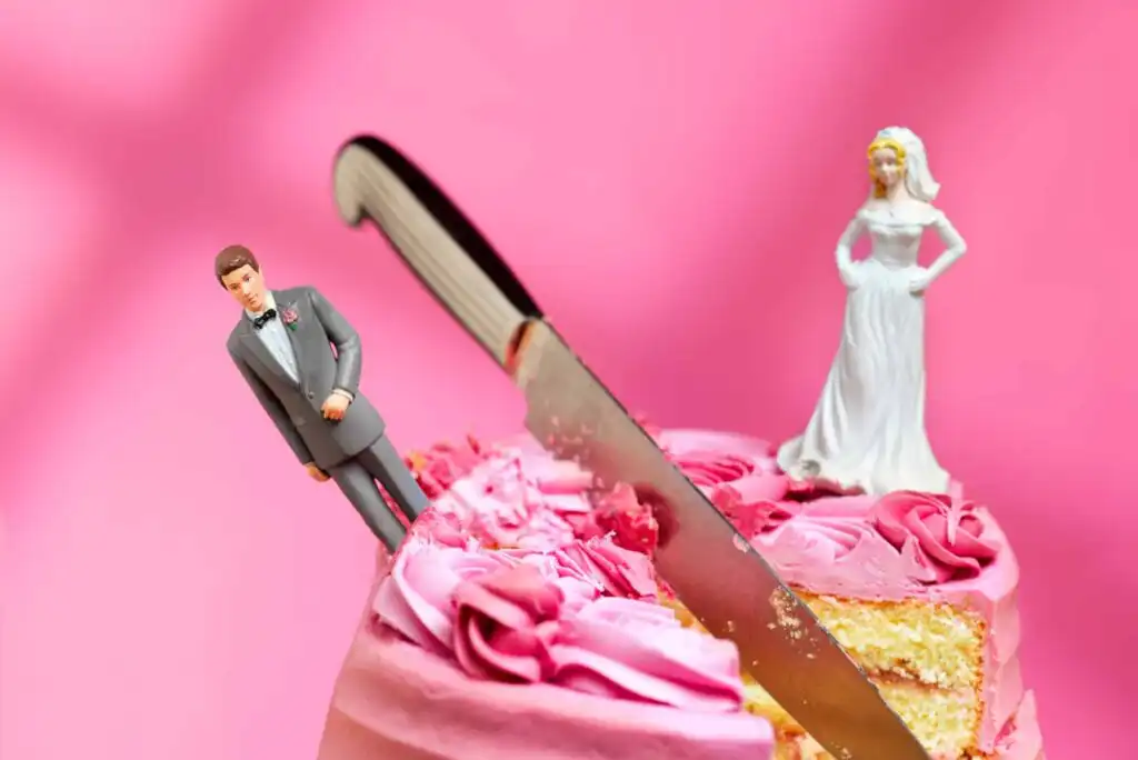 A wedding cake with pink frosting shows a knife cutting between bride and groom figurines placed on top, against a pink background.