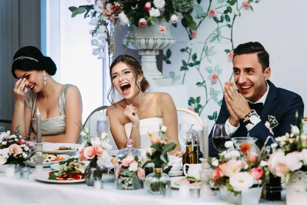 Three people in formal attire sit at a decorated table, laughing and smiling during a celebration. Floral arrangements and elegant table settings surround them, creating a joyful, festive atmosphere.