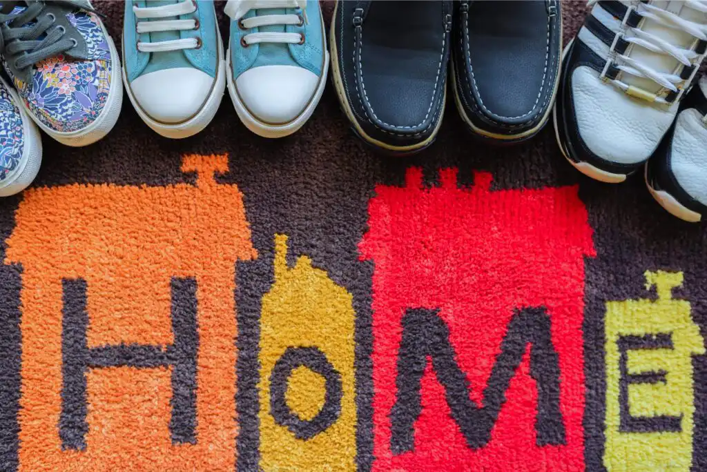 Four pairs of shoes are lined up on a colorful doormat that spells HOME with stylized house shapes forming each letter. The shoes suggest people entering or gathering at home.