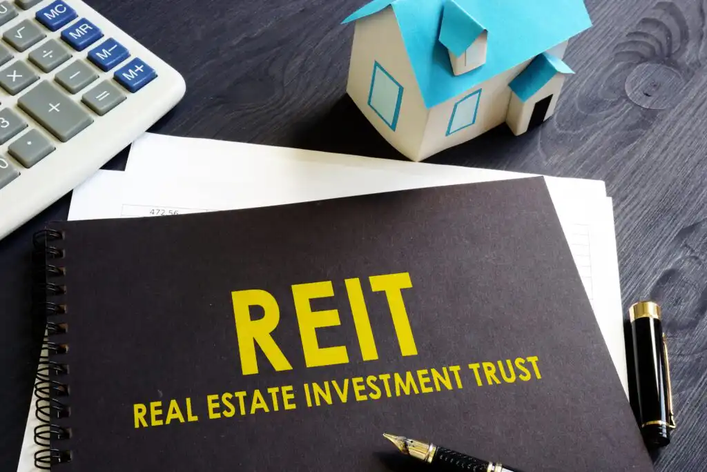 A notebook labeled REIT Real Estate Investment Trust sits on a desk beside a pen, calculator, papers, and a small model house with a blue roof.